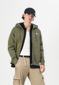 Sweatshirt com fecho éclair na cor verde-azeitona, com um bolso frontal e cordões, usado em camadas sobre uma camisa preta de botão e uma t-shirt branca, combinado com calças de trabalho beiges.