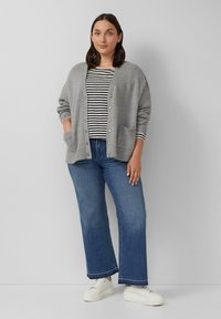 Grijze cardigan met voorzakken, ronde halslijn en knopen over een gestreept shirt met lange mouwen. Gecombineerd met blauwe wijde jeans en witte sneakers.