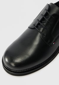 Μαύρο δερμάτινο παπούτσι με λεία επιφάνεια, στρογγυλή μύτη και σχέδιο με έξι τρύπες για κορδόνια. Διακριτή λεπτομέρεια ραψίματος κατά μήκος της σόλας.