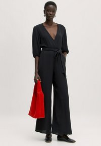 Zwarte, geribbelde jumpsuit met een diepe V-hals, een cinched taille en een wijd broekontwerp. Gecombineerd met een felrode suède tas. Zwarte schoenen.