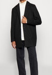 Černé blazer s texturou, dvouknoflíkový, střih na míru, nošený přes bílý rolák. Rukávy jsou ohrnuté tak, aby byl vidět vnitřní materiál.