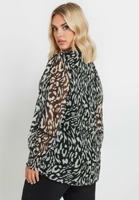 Blusa a righe in bianco e nero con maniche trasparenti marroni, caratterizzata da unfit rilassato e polsini arricciati. Il tessuto presenta una texture vorticosa.