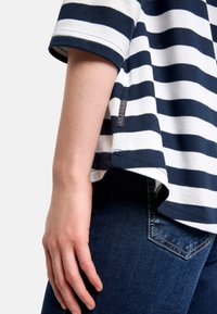 Gestreiftes T-Shirt in Marineblau und Weiß, aus Baumwolle, mit lockerem Schnitt, hochgekrempelten Ärmeln und einem Etikett am Saum. Kombiniert mit dunkelblauen Jeans.