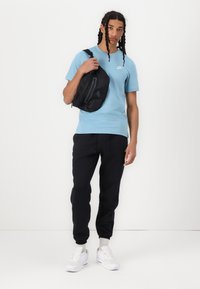 Ljusblå bomulls-T-shirt med en liten vit logotyp, svart axelremsväska med grå detaljer, svarta joggers och vita sneakers med synlig luftdämpning.