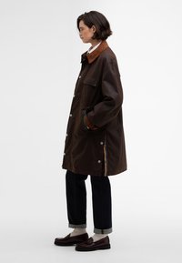 Barna külső dzseki bordás gallérral, patentgombokkal, oldalzsebekkel és színes csíkos részletekkel az varrásokon. Sötét farmerrel és cipőkkel párosítva.