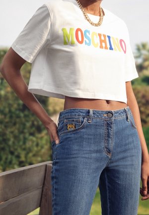 Witte cropped t-shirt met multicolored "MOSCHINO" lettertype, gecombineerd met blauwe denim jeans met een berenpatch op de voorzak.