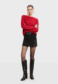 Roter, glitzernder Pullover kombiniert mit einem schwarzen Minirock, der einen seitlichen Reißverschluss und einen Gürtel hat. Sie trägt kniehohe, glänzende schwarze Stiefel.