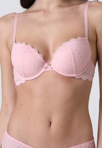Reggiseno in pizzo rosa con bordi a scallop e spalline sottili indossato da una donna su uno sfondo neutro, che mostra il ventre e la parte superiore dei fianchi.