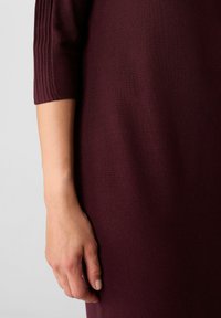 Braccio sinistro e mano poggiati accanto al corpo, indossando un indumento lavorato a maglia color bordeaux scuro con maniche a tre quarti a coste.