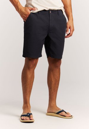 Man draagt zwarte knielange shorts, beige en marineblauwe teenslippers, staat met één hand in de zak tegen een effen achtergrond.
