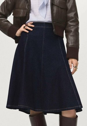 Persoon draagt een donkerbruine leren jas, lichtblauwe blouse, marineblauwe A-lijn rok en bruine kniehoge laarzen, met ringen aan beide handen.