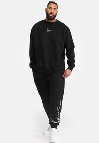 Conjunto de sudadera y pantalones de chándal negros, confeccionado con tela suave. Presenta un logotipo en blanco en el pecho y en la pierna. Completado con zapatillas blancas.