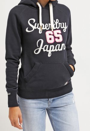 Osoba ubrana w czarną bluzę z kapturem Superdry z białymi sznurkami, numer '65' w kolorze różowym oraz jasnoniebieskie dżinsy z przetarciami.