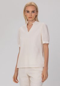Blouse blanche à manches courtes perforée avec un décolleté en V, présentant des détails de col volantés et une coupe ample et décontractée. Texture de tissu lisse.
