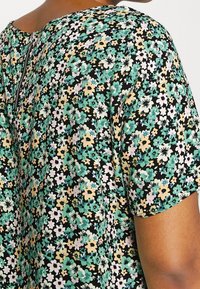 Blommig topp med korta ärmar, med en svart bakgrund och ett mönster av gröna, gula och rosa blommor. Inkluderar en dragkedjedetalj bak.