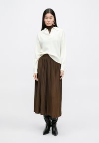 Λευκό πουλόβερ από μαλλί με V-λαιμό και φουσκωτά μανίκια, συνδυασμένο με πλισέ καφέ μάξι φούστα και μαύρες μυτερές μπότες μέχρι το γόνατο.