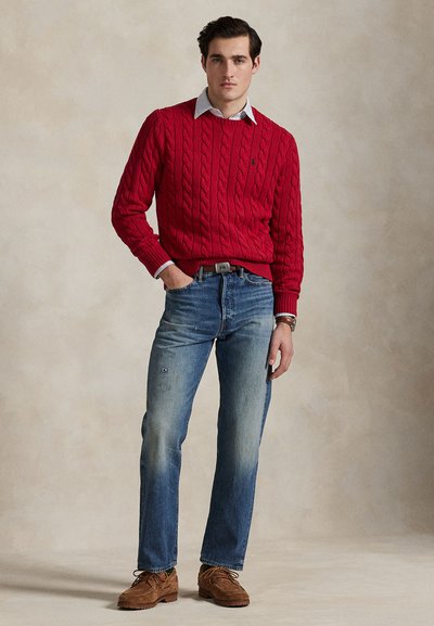 Giovane uomo che indossa un maglione rosso a trecce, jeans azzurri e scarpe marroni con lacci, in piedi davanti a uno sfondo beige.