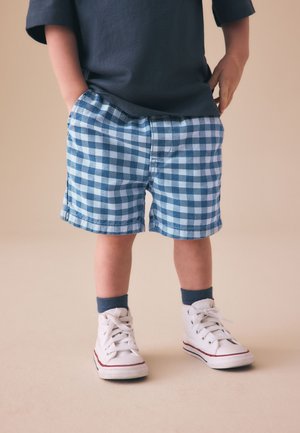 Barn iført blå ternede shorts, sorte sokker, hvide høje sneakers og en mørk navy trøje med hænderne i lommerne.