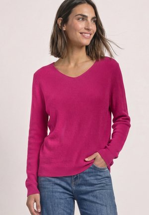 Femme souriante, portant un pull à col en V à manches longues rose vif et un jean bleu, avec une main dans la poche sur un fond uni.