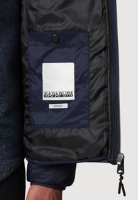 Intérieur de la veste Napa Puri avec doublure bleu marine, arborant un écusson logo blanc et une poche à bouton pression noire. Tissu lisse et léger.