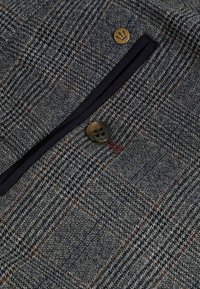 Tissu en mélange de laine dans un motif pied-de-poule bleu marine et gris, présentant un bouton marron et une poche zippée avec un contour noir.