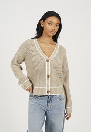 Gilet en laine beige avec un col en V, rehaussé d'une bordure blanche, comportant quatre boutons ronds et des manches longues. Porté avec un jean bleu clair.