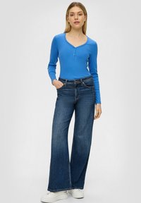 Langsleeve, geribbelde blauwe top met drie knopen, gecombineerd met wijde, donkerblauwe jeans en witte platformsneakers. Eenvoudig ontwerp, casual uitstraling.