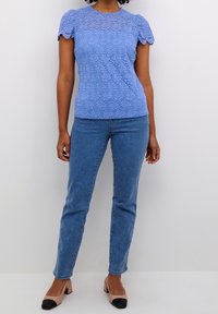 Haut en dentelle bleu clair avec motif floral, manches courtes bouffantes, associé à un jean denim bleu et des talons beiges avec embouts noirs.