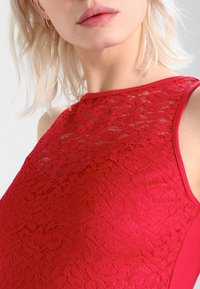 Czerwona sukienka z koronkową nakładką na górze, z gładkim, jednolitym czerwonym materiałem po bokach oraz wysokim dekoltem.