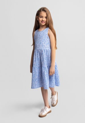 Młoda dziewczyna stojąca, ubrana w bezrękawnikową niebieską sukienkę w kwiaty i białe trampki, delikatnie uśmiechająca się na tle jednolitego jasnego tła.