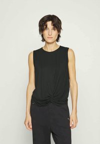 Woman with short dark hair wearing a sleeveless black knotted top and black pants, standing against a plain light background.