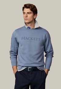 Giovane uomo con le mani in tasca che indossa una felpa blu Hackett London sopra una camicia con colletto, su uno sfondo semplice.