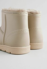 Beige ankelstøvler med blødt, fluffy foer, glat læderlignende materiale og stikninger, der har en tekstureret gummisål.