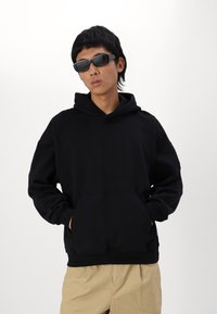 Schwarzer Kapuzenpullover mit langen Ärmeln, Fronttasche und gerippten Bündchen. Getragen mit hellbeigen Shorts und schwarzen Sonnenbrillen.
