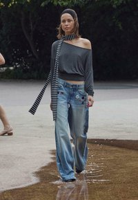 Tânără mergând pe trotuar umed, purtând o bluză în dungi cu umerii goi, cu o eșarfă asortată și blugi cu picioare largi decorați cu petice și broșe.