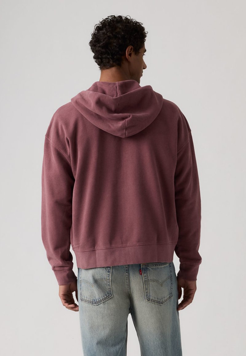 Man draagt een hoodie in stoffig roze en lichtblauwe spijkerbroek, gezien van achteren tegen een effen lichte achtergrond.