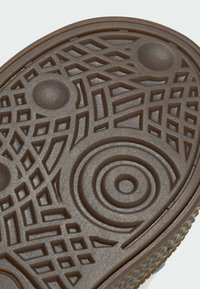 Hnědá gumová podrážka s komplikovanými texturovanými vzory, včetně kruhů, diamantů a čar pro trakci a přilnavost.