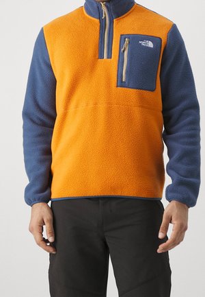 Mann trägt einen Fleecepullover mit leuchtend orangefarbenem Körper, dunkelblauen Ärmeln und einer Reißverschlusstasche auf der Brust mit dem Aufdruck The North Face.
