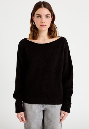 Giovane donna con capelli castani alla lunghezza delle spalle, che indossa orecchini a cerchio dorati, un maglione nero ampio e jeans grigio chiaro su sfondo bianco.