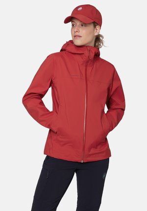 Femme portant une veste à capuche rouge et une casquette rouge assortie, mains dans les poches de la veste, regardant sur le côté sur un fond uni.