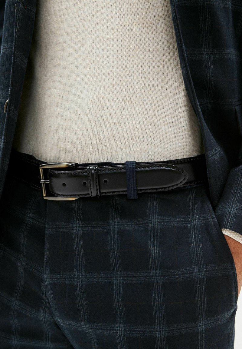 Ceinture en cuir noir avec une boucle en argent, portée avec un costume à carreaux bleu marine. Texture lisse, complétée par un pull de couleur claire en dessous.