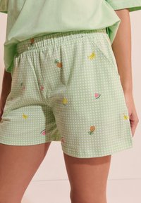 Pantalones cortos verde claro con patrón de cuadros blancos y pequeños estampados de frutas que incluyen piñas, plátanos, cerezas y rodajas de sandía.