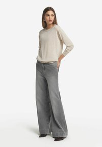 Hellbeige Langarmshirt kombiniert mit hochtaillierten, weit geschnittenen grauen Jeans. Das Outfit zeichnet sich durch eine strukturierte, lockere Passform und ein dezentes Muster am Saum aus.