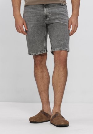 Mand iført grå denimshorts og brune ruskind slip-on sko, stående mod en ensfarvet lys baggrund.