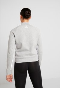 Grå sweatshirt med høj krave, ribbede manchetter og kant. Har hvide sidestriber og et Adidas-logo på bagsiden. Glat stofstruktur.