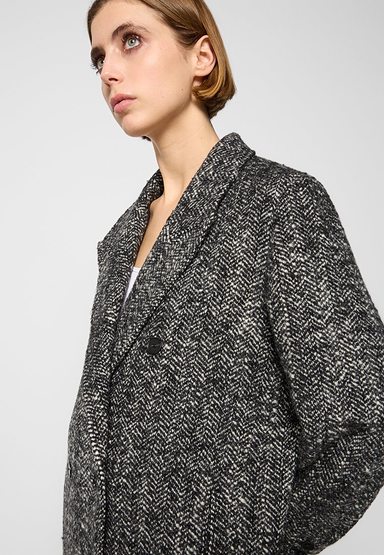 Jonge vrouw met kort haar die een zwart-witte gebreide wollen jas draagt met gekartelde revers en een enkele zwarte knoop.