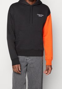 Sudadera negra con una manga naranja, hecha de una mezcla de algodón, que presenta un bolsillo tipo canguro y el logo de Calvin Klein Jeans en blanco en el pecho.