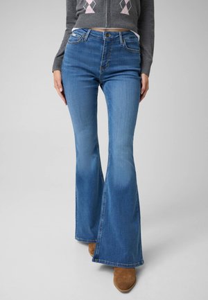 Kvinde iført blå højhælede flare-jeans, grå cropped sweater og brune spidse støvler, står mod en ensfarvet baggrund.