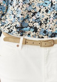 Chemise fleurie en noir, bleu et crème ; ceinture en daim beige clair avec boucle rectangulaire en or ; pantalon en denim blanc avec une poche avant.