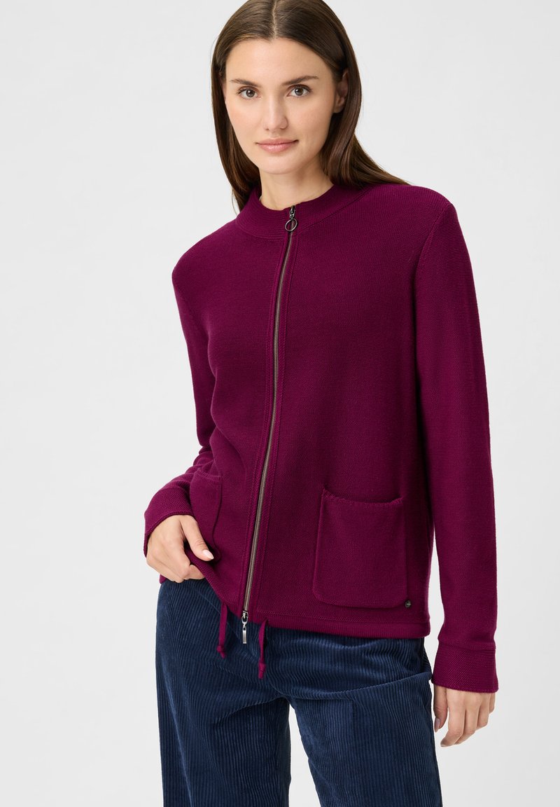 Magenta zip-up cardigan with a round neck, featuring a front pocket and ribbed cuffs. Worn over navy corduroy pants.